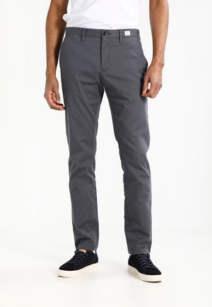 Mężczyzna ubrany w szare dopasowane spodnie, białą koszulkę i czarne trampki z białą podeszwą, stojący na tle jednolitego białego tła.