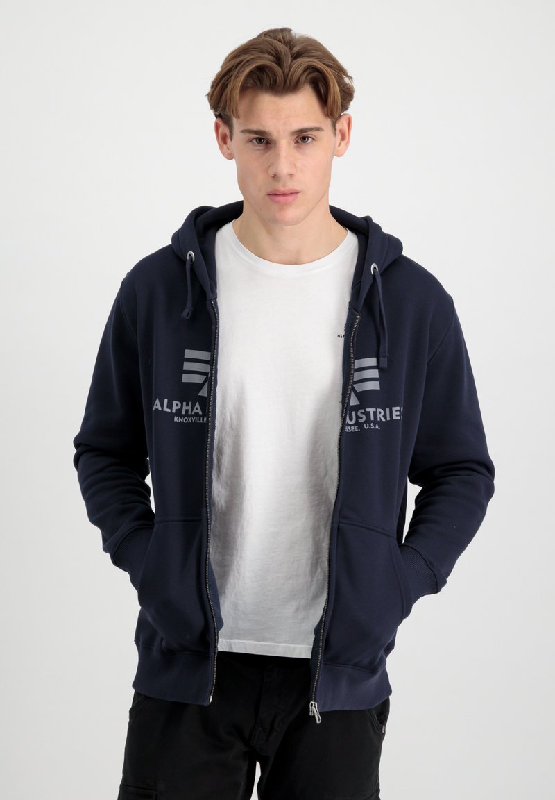 Mornarska zip-up dukserica s kapuljačom na vezanje, prednjim džepovima i otisnutim logotipom. Nosena preko bijele majice, s mekim materijalom i ležernim krojem.