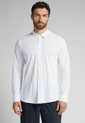 Langärmliges weißes Hemd mit button-down Kragen, glatter Textur und figurnahem Schnitt. In Kombination mit dunkler Hose, minimaler Details.