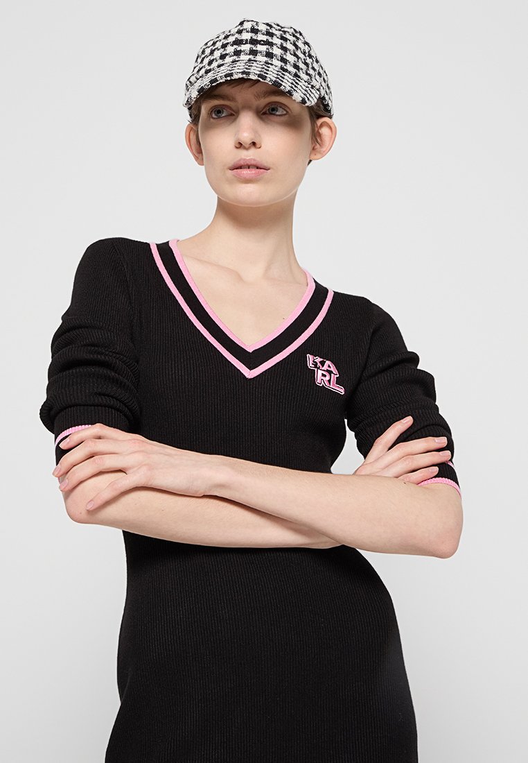 Schwarzer gerippter Pullover mit V-Ausschnitt, akzentuiert mit pinken Besatz und einem Logo. Das Model trägt eine schwarz-weiße karierte Mütze.