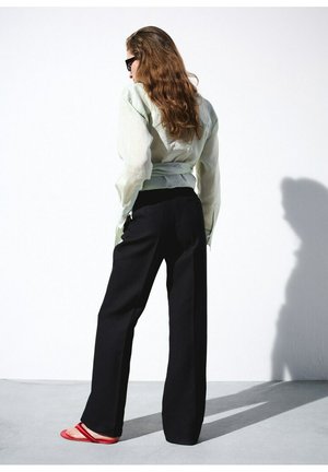 Mujer con cabello largo que lleva pantalones negros, blusa transparente de color claro, gafas de sol negras y sandalias rojas, de pie contra una pared blanca.