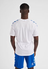 Witte sportshirt met blauwe accenten op de schouders, gecombineerd met blauwe shorts met een wit chevronpatroon. Glad, lichtgewicht materiaal.