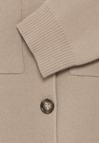 Maglione beige con un design in maglia texturizzata, polsini a coste e un bottone marrone rotondo nella parte anteriore.