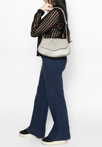 Borsa a tracolla in pelle grigia con bordi superiori ondulati, spallacci e una texture morbida, abbinata a jeans svasati blu scuro e sneaker nere glitterate.