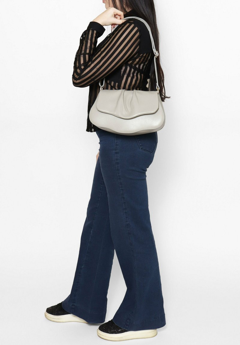 Borsa a tracolla in pelle grigia con bordi superiori ondulati, spallacci e una texture morbida, abbinata a jeans svasati blu scuro e sneaker nere glitterate.