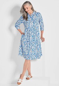 Blau-weiß gemustertes Hemdkleid mit durchgehender Knopfleiste, dreiviertel Ärmeln, Taillengürtel und seitlichen Taschen. Tragend mit weißen Sandalen.