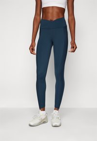 Sötétkék, magas derekú leggings sima textúrával és kontrasztos oldalsó panelek nélkül. Fehér sportcipővel és fehér bokazoknival viselve.