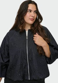 Chaqueta negra corta con un patrón floral texturizado, que presenta un cierre frontal y mangas anchas. El material parece brillante y suave.