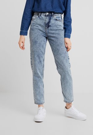 Personne portant un jean droit taille haute, délavé clair, avec un haut à manches longues bleu foncé et des baskets blanches, debout.