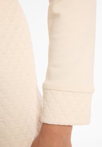 Hellbeige Langarmkleid mit einem strukturierten Wabenmuster am Saum. Glatte Ärmelstoffe, taillierte Manschettenverarbeitung.