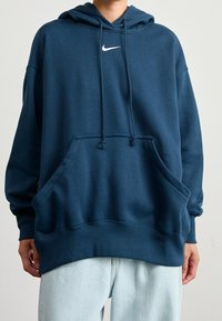 Persona vestida con una sudadera oscura de Nike con bolsillo frontal y jeans de un tono azul claro, de pie frente a un fondo neutro.