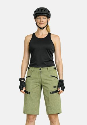 Femme portant un casque noir, un haut sans manches noir, un short de vélo vert et des gants noirs, debout devant un fond uni.