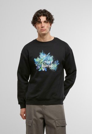 Črna sweatshirt s pisanim grafičnim dizajnom, ki vključuje modre in zelene akcentne barve, okroglo ovratnico in rebraste manšete. Nosijo jo s sivimi cargo hlačami.