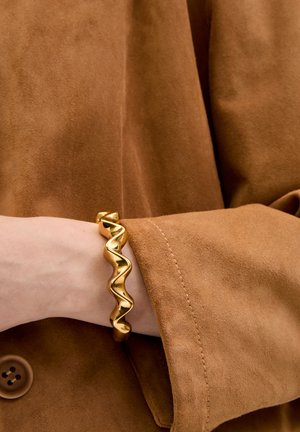 Gold wave-patterned metal bracelet worn on a wrist, contrasting with a soft brown suede jacket visible in the background.