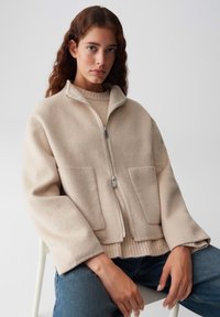 Donna con lunghi capelli mossi indossa una giacca di lana beige con zip sopra un maglione beige e jeans blu, seduta su una sedia bianca contro uno sfondo neutro.