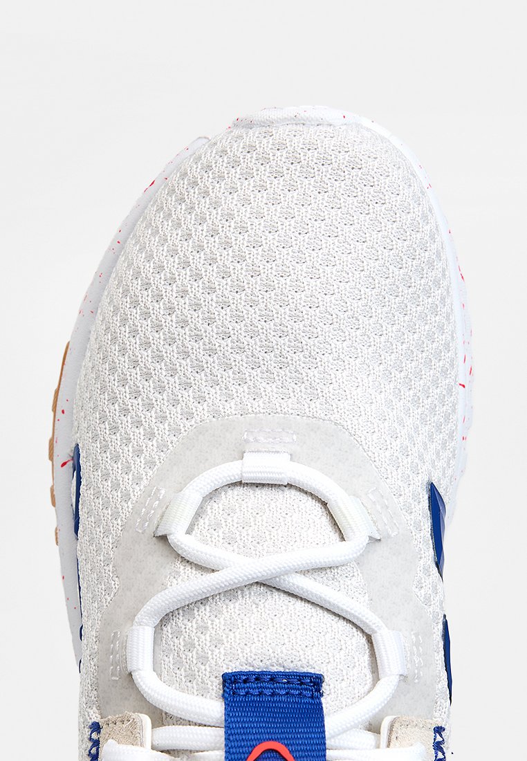 Zapatilla deportiva de malla blanca con detalles en ante gris, logotipos en azul y diseño intrincado de cordones. Presenta una superficie texturizada y suela de goma.