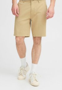 Shorts beiges à coupe droite, fabriqués en tissu léger. Présentent des poches avant et une ceinture plate. Associés à des baskets blanches et des chaussettes.
