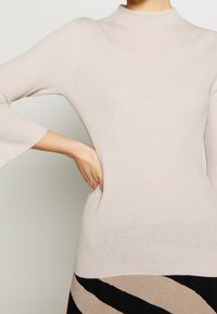 Donna che indossa un top a maniche lunghe beige a costine con polsini svasati, mano sulla vita, abbinato a una gonna con strisce diagonali nere e beige.
