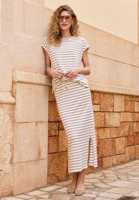 Femme aux cheveux bouclés portant un haut sans manches rayé et une jupe longue assortie avec une fente latérale, debout contre un mur en pierre.