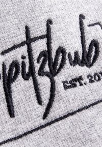 Grau gestrickter Stoff mit auffälligem schwarzen gesticktem Text in stilisierten Buchstaben und einem festgelegten Datum, vor einem strukturierten Hintergrund.