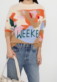 Sweter dziany z wielokolorowym abstrakcyjnym wzorem i napisem „WEEKEND” w zielonym kolorze, zestawiony z niebieskimi dżinsami z wysokim stanem oraz teksturowaną torbą.