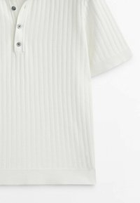 Chemise blanche à manches courtes en tricot côtelé avec trois boutons foncés sur une encolure à patte, présentée à plat sur fond blanc.