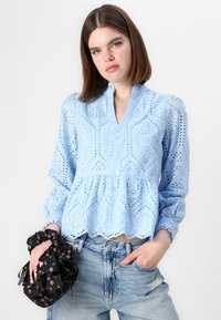 Blusa azzurro chiaro in pizzo con maniche a sbuffo e orlo peplum, con ritagli floreali e una finitura morbida e texturizzata. Scrunchie nero con motivo in mano.
