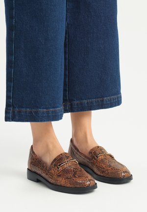Bruine loafers met slangenpatroon, met een ronde neus, platte zool en goudkleurige accenten, gecombineerd met donkerblauwe wijdvallende denim.