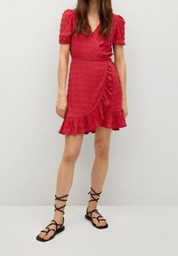 Rotes Wickelkleid mit kurzen Puffärmeln, strukturiertem Stoff, gerafftem Saum und seitlichem Bindeband. Getragen mit schwarzen, schnürbaren Sandalen.