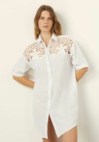 Camicia bianca con bottoni, caratterizzata da un motivo a crochet di pizzo sulle spalle. Presenta maniche arrotolate e un orlo leggermente allungato sui lati.