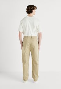 Pantaloni din bumbac bej cu o croială lejeră. Spatele dispune de două buzunare cu nasturi și bucle pentru curea. Împreună cu o cămașă cu mânecă scurtă de culoare deschisă.
