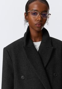 Dunkelgrauer, doppelt knöpfbarer Mantel aus strukturiertem Stoff, mit großem Kragen und zwei schwarzen Knöpfen. Modell trägt eine Brille, neutraler Hintergrund.