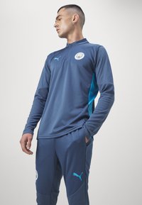 Completo da allenamento Puma di colore blu scuro con accenti turchesi, dotato di colletto con zip, tessuto testurizzato e logo del team sul petto e sui pantaloni.