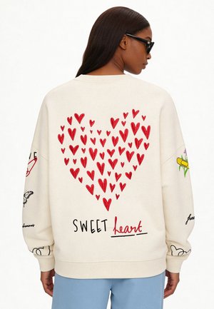 Persoon draagt een crèmekleurige trui met rode harten die een groot hart vormen en de tekst "SWEET heart" op de achterkant, staand tegen een effen achtergrond.