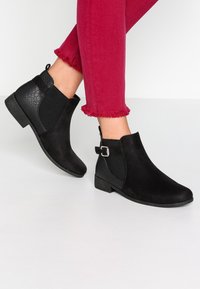 Botas negras hasta el tobillo con un panel texturizado negro, una parte delantera suave y un tacón pequeño. Los detalles incluyen un acento de hebilla en el costado.