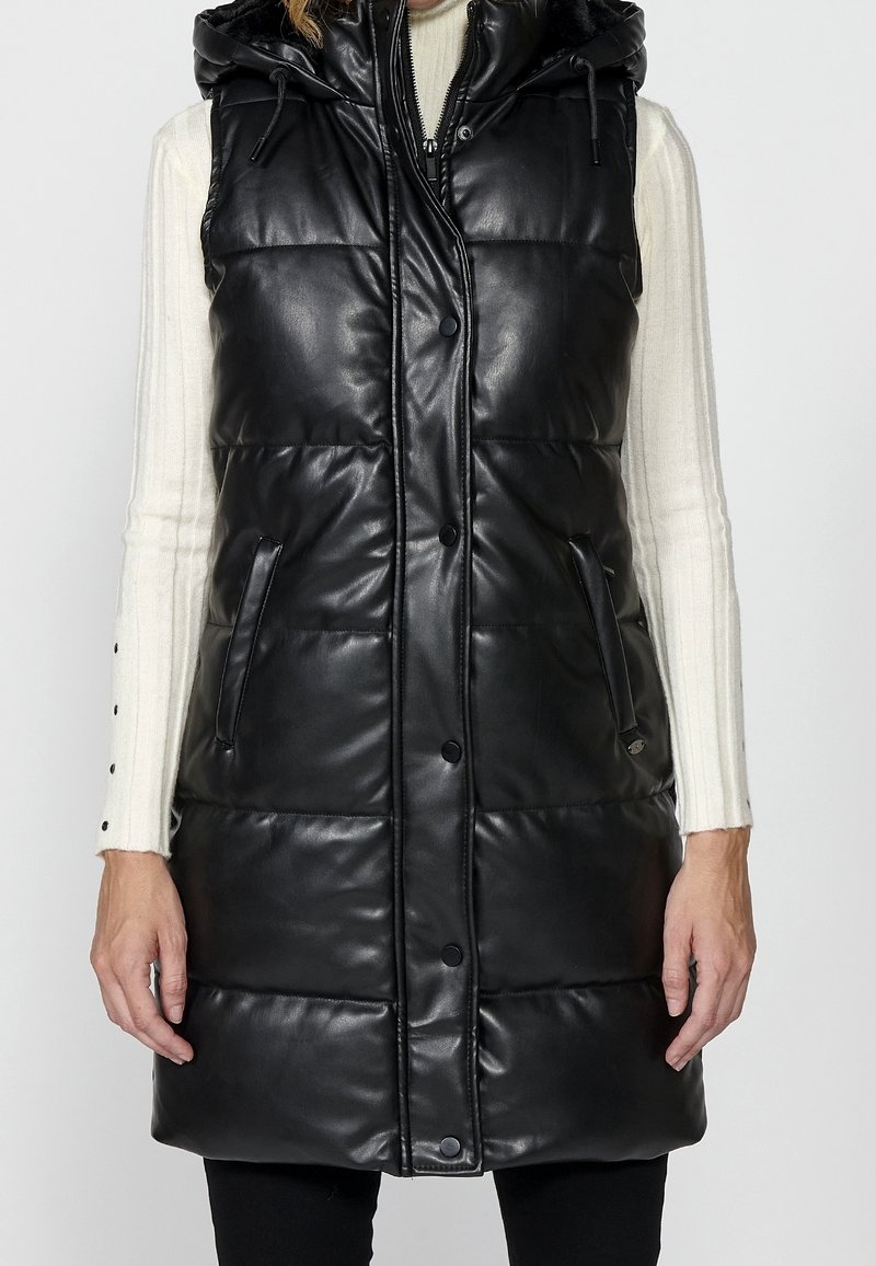 Femme portant une veste matelassée sans manches en similicuir noir avec des boutons à l'avant et une capuche par-dessus un pull blanc côtelé.
