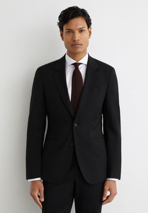 Sötét hajú férfi fekete öltönyt, fehér inget és texturált sötétbarna nyakkendőt viselve, egy fehér falnak támaszkodik.