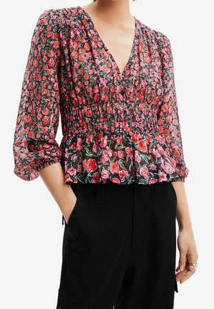 Femme portant un chemisier noir transparent avec un imprimé floral rouge et rose, encolure en V, taille froncée, manches trois-quarts bouffantes et pantalon noir.