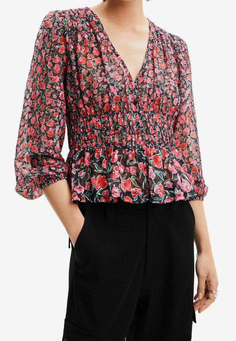 Femme portant un chemisier noir transparent avec un imprimé floral rouge et rose, encolure en V, taille froncée, manches trois-quarts bouffantes et pantalon noir.