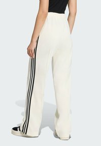 Krémové teplákové nohavice s voľným strihom, ktoré majú tri čierne vertikálne prúžky po stranách a elastický pás.