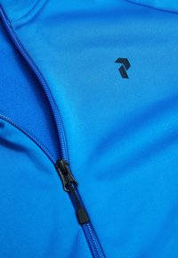 Blå zip-up jakke lavet af glat, letvægtsstof. Har et sort logo og kontrasterende lynlåsdetaljer. Ingen synlige mønstre eller accenter.