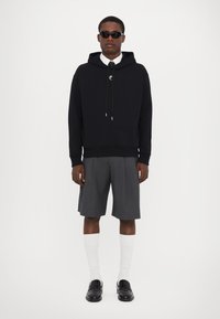 Hanorac negru cu șnururi, pantaloni scurți gri, șosete albe până la glezne și loafers negri. Modelul poartă ochelari de soare cu un design curat și minimalist.