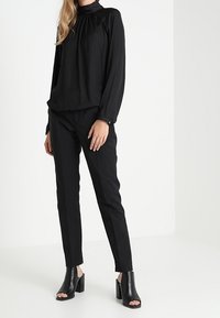 Femme portant un blouse noir à col montant, des pantalons noirs ajustés, et des bottines à talons ouverts noires se tenant sur un sol blanc.