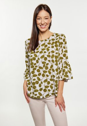 Donna sorridente che indossa una camicetta bianca con motivi di foglie verdi e pantaloni beige, in piedi davanti a uno sfondo bianco semplice.
