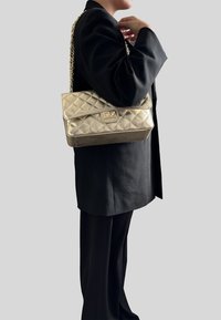 Złota pikowana torebka na łańcuszku, z klapą i prostokątnym metalowym akcentem. Gładka tekstura i zorganizowany design.