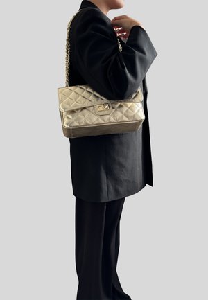 Bolso de mano acolchado dorado con correa de cadena, que cuenta con un cierre de solapa y un acento metálico rectangular. Textura suave y diseño estructurado.