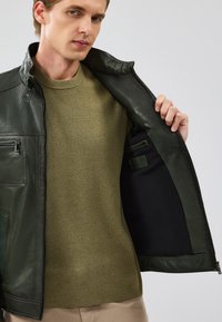 Giacca di pelle verde scuro con hardware argento, tasca con cerniera e finitura testurizzata, indossata sopra un maglione verde oliva. Fodera interna nera.