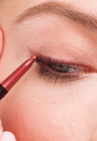 Crayon eye-liner rouge appliquant la couleur le long de la ligne des cils d'un œil. Présente des cils foncés et une texture de peau lisse autour de la zone des yeux.