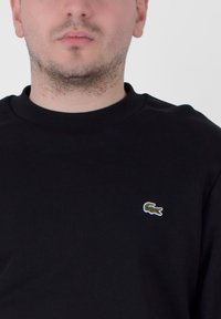 Black cotton sweatshirt featuring a crew neck and a small green crocodile logo on the left chest. Smooth texture, simple design.