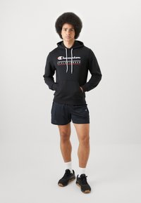 Svart hettegenser med hvite snorer har et grafisk "Champion"-logo i rødt og hvitt. Kombinert med svarte shorts og svarte joggesko.